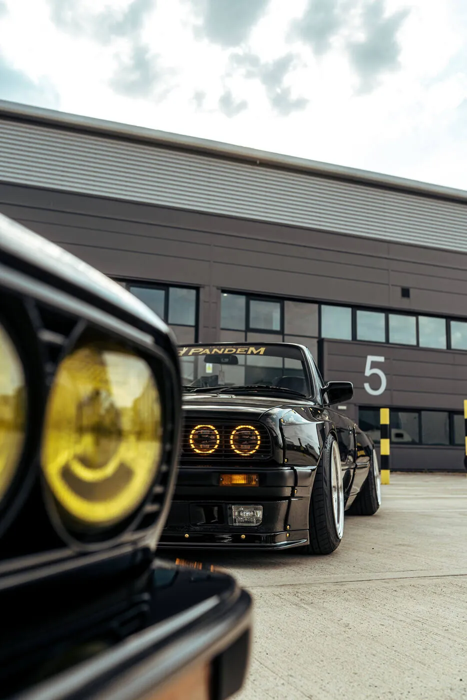 Yellow headlight of a black E30 in the foreground, more cars parked beyond