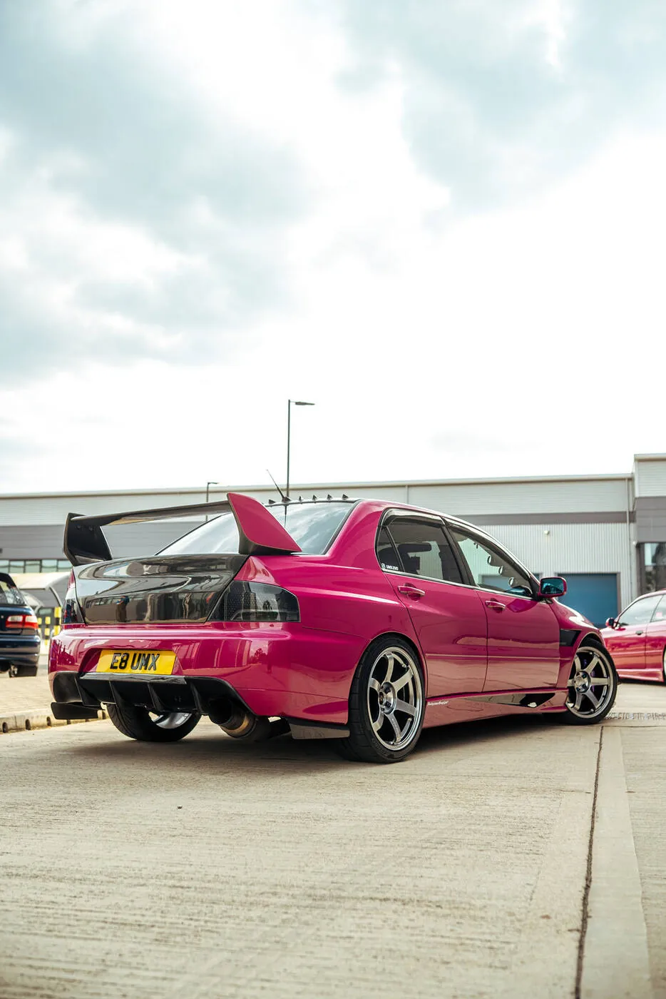 Magenta Lancer Evolution from the rear, its wing against the sky on an industrial estate